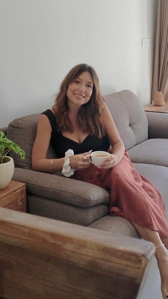 Elaine Palmer sitting on a sofa holding a cup, plant and window behind her